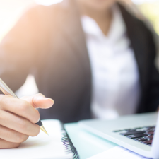 Closeup of laptop and pen writing in notebook with a person in the background