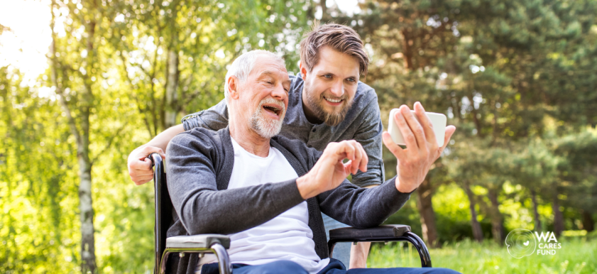 older man in wheelchair taking selfie with young adult standing behind him