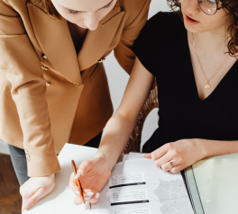 two women at a desk across from each other with a notebook open and pen